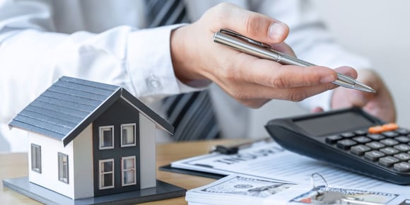 A hand holding a pen with a calculator, money, and a small model of a home.