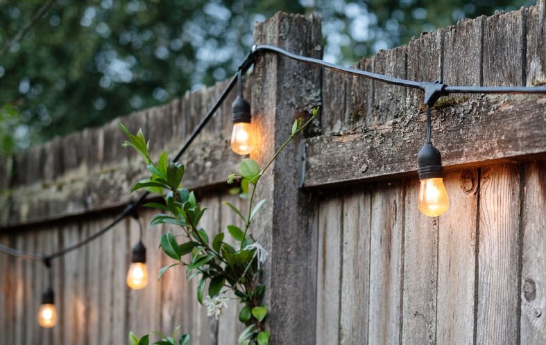 A wooden fence has string lights hanging from it with the lights on in the dusk.