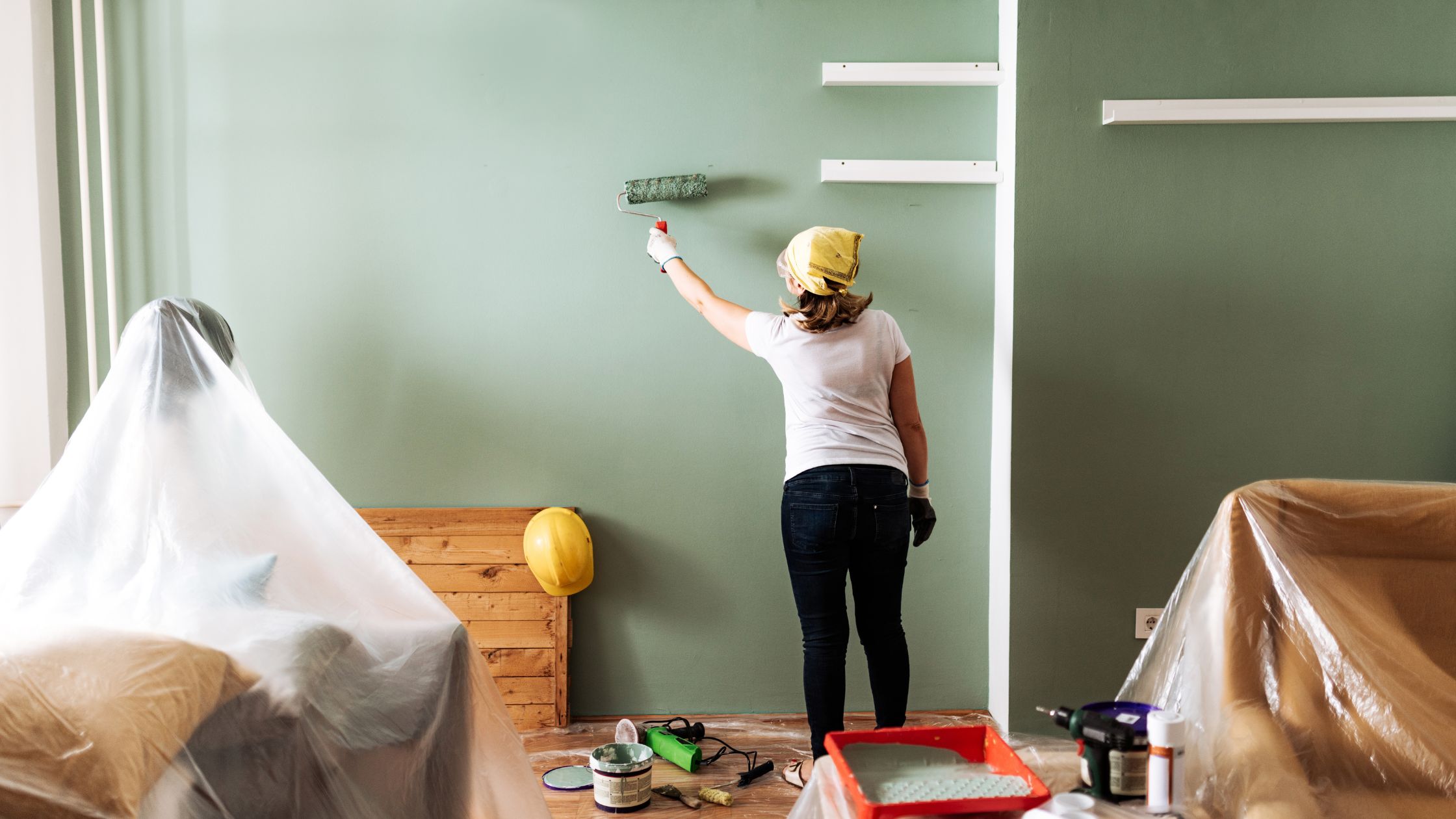 A woman paints a wall green in her home and is surrounded by other DIY home improvement tools liks hammers and a helmet.
